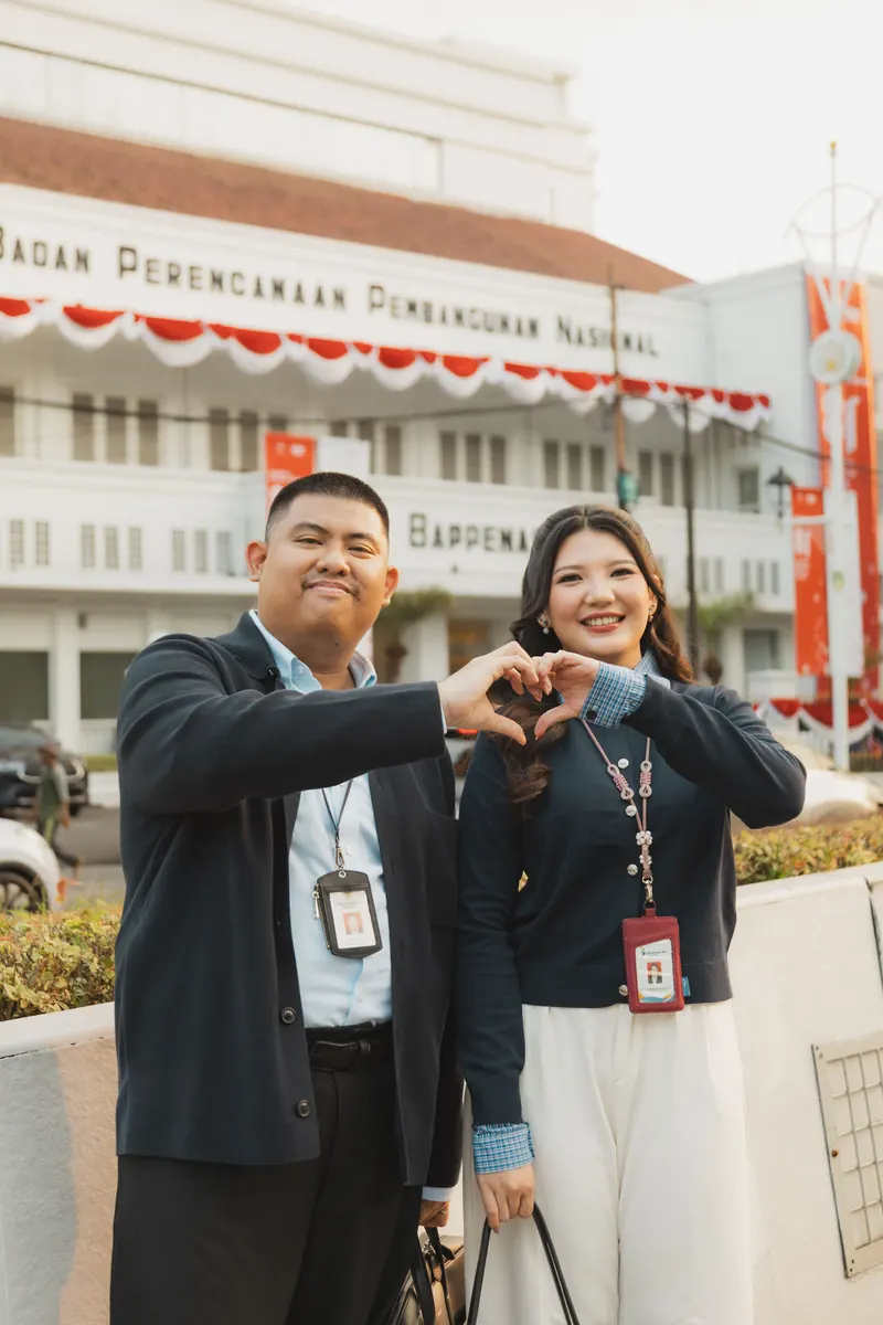 Rafli dan Dita berbaju kantor dengan tanda pengenal, membentuk hati dengan tangan di depan gedung Bappenas