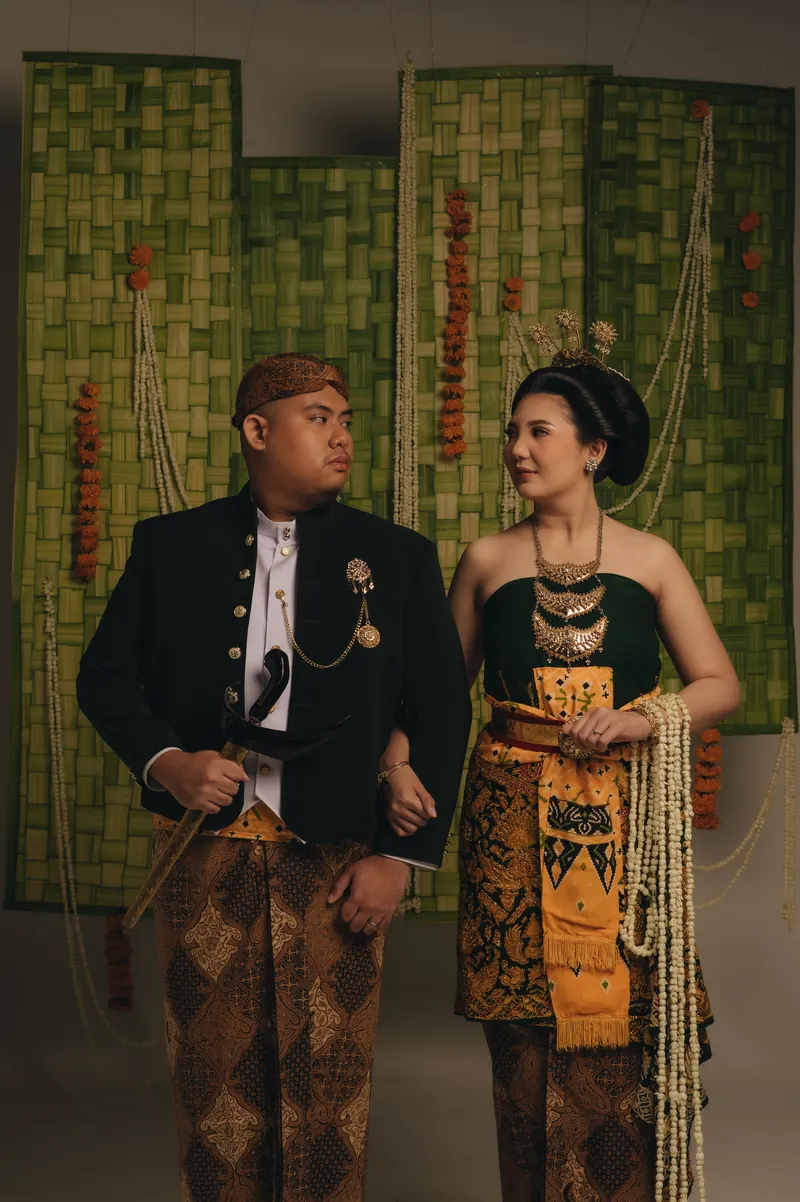 Rafli dan Dita berdampingan dalam busana Jawa, dengan latar anyaman hijau