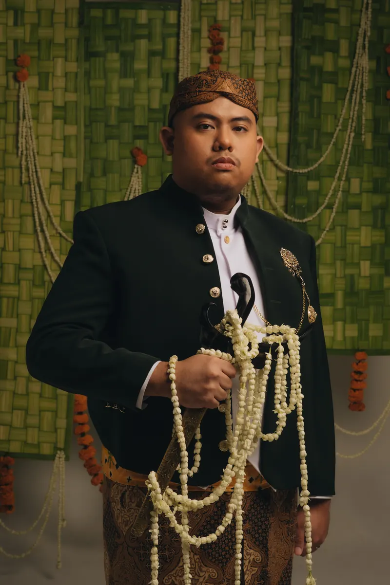 Rafli dalam beskap hijau tua dan batik, memegang untaian melati