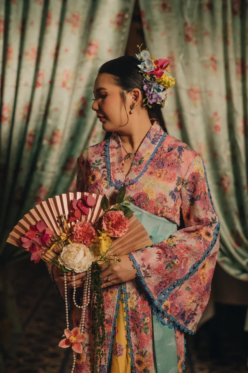 Dita dalam kebaya merah muda bermotif bunga, memegang kipas kertas dan buket